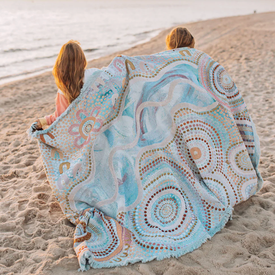 Two people sitting on a sandy beach with a large decorative blanket.