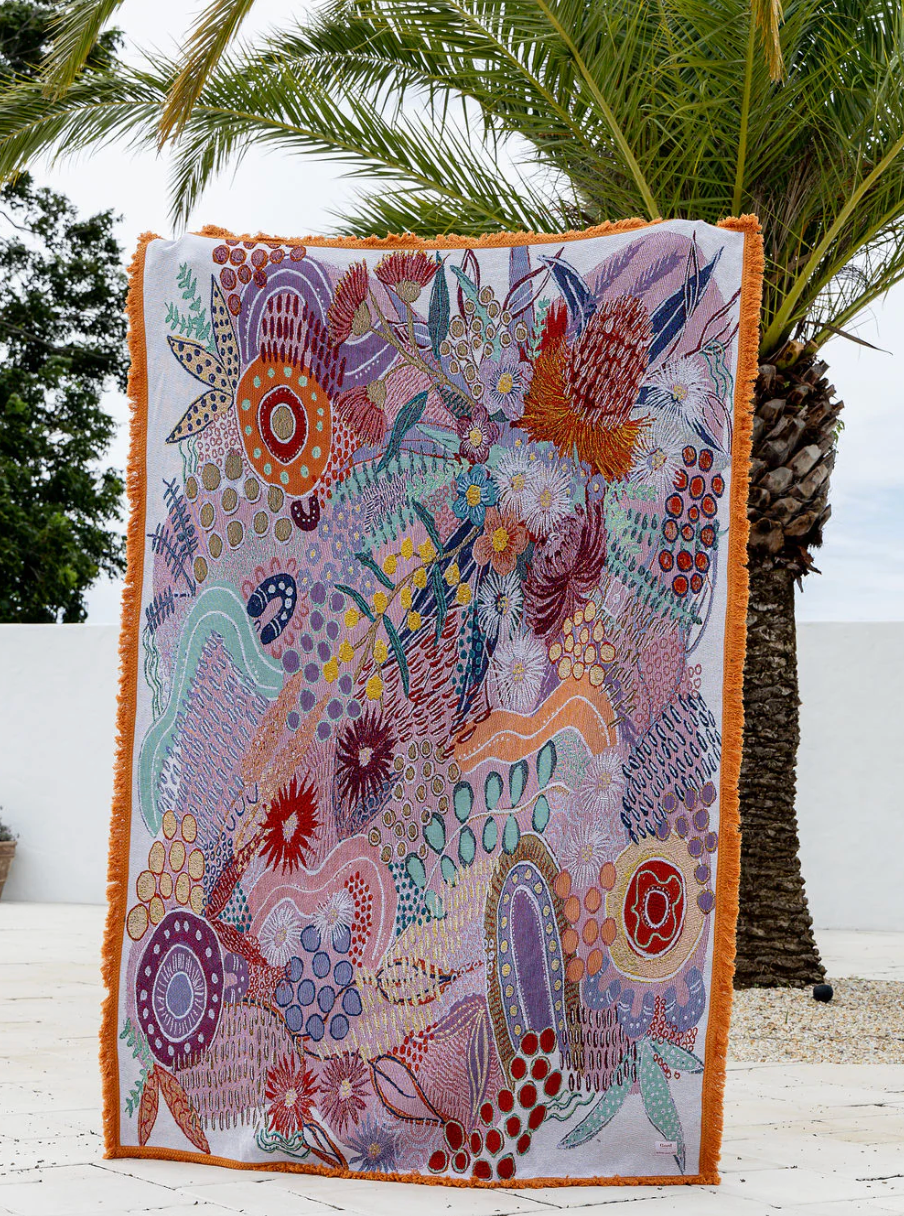 Colorful abstract patterned blanket with fringe on a wooden floor against a palm tree.