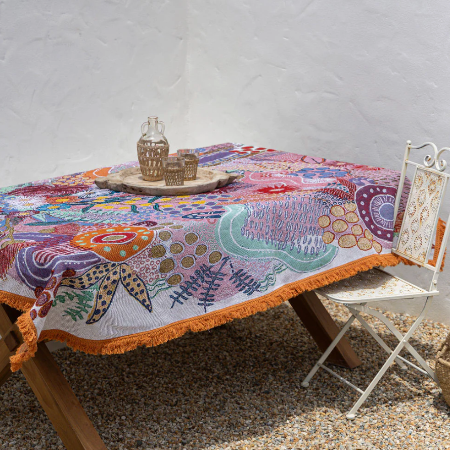 Colorful patterned tablecloth on a wooden table with a white wall and greenery in the background.