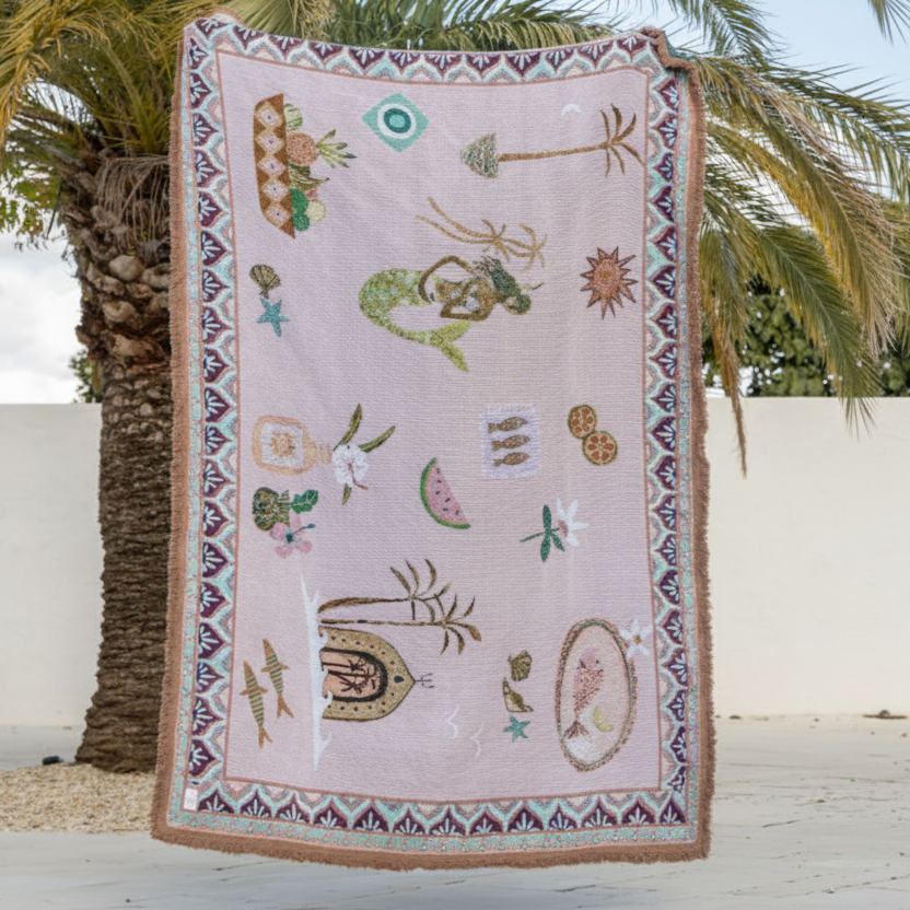 Decorative blanket with mermaid and shell patterns on a wooden surface with palm trees in the background
